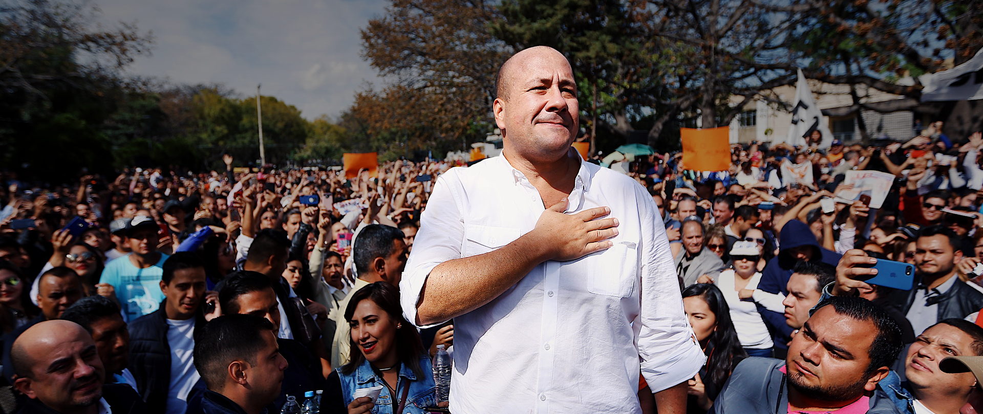 Foto de perfil del gobernador hablando en un podium con micrófono, al fondo una pared de madera tallada con rectangulos alargados